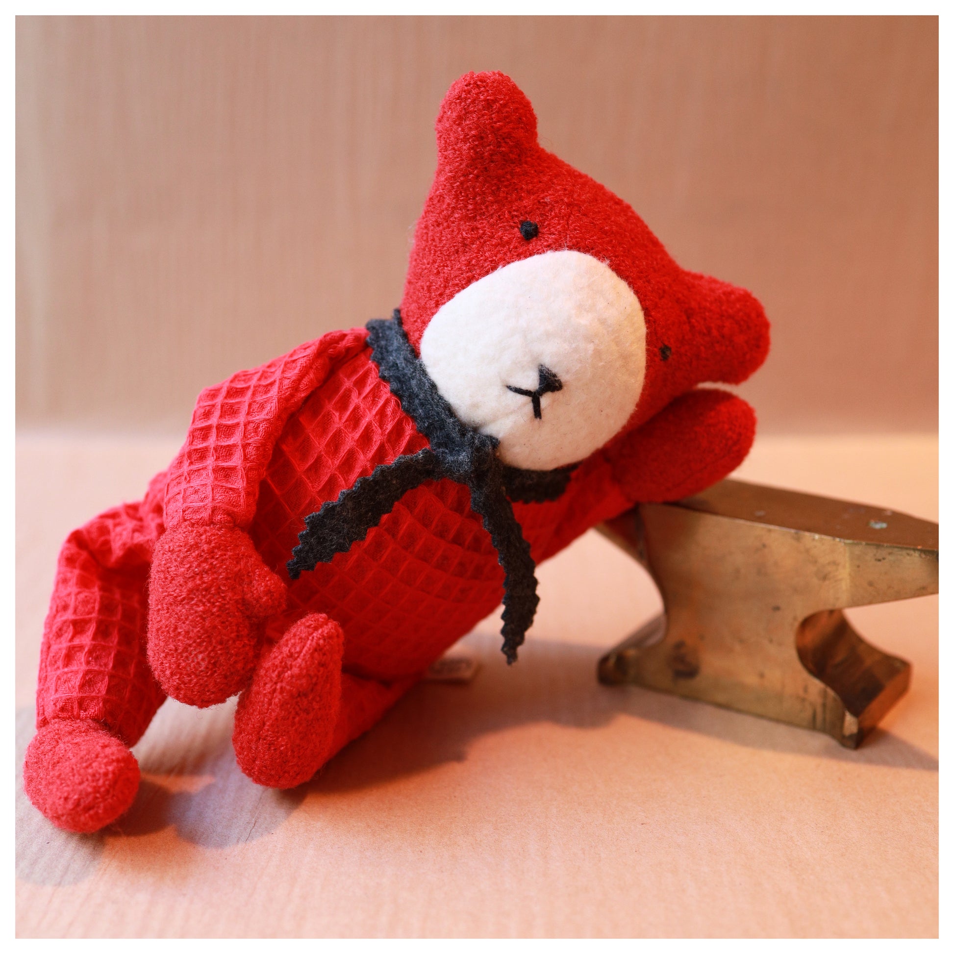 Red teddy bear with a white muzzle leaning on a brass anvil and wearing a grey wool scarf