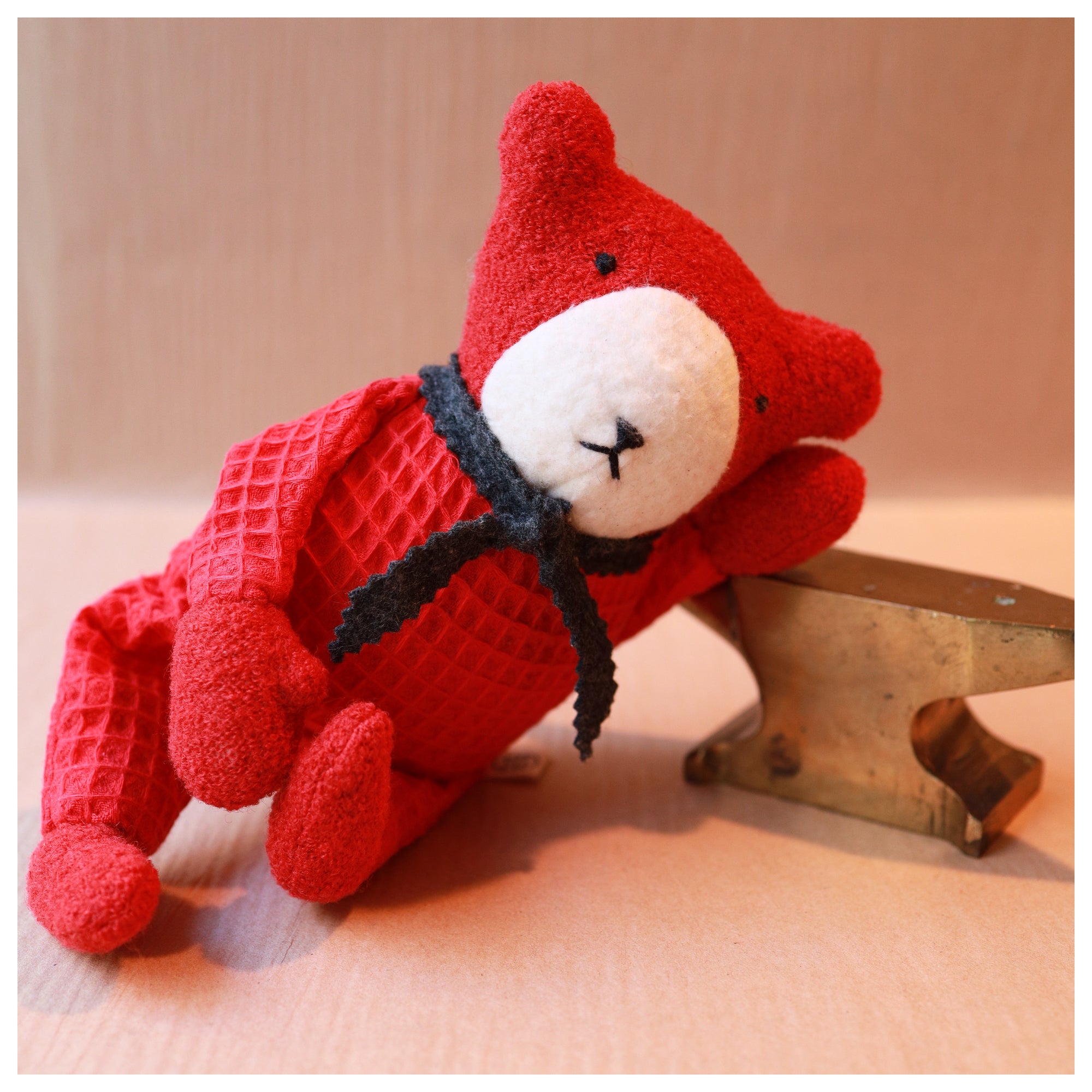 Red teddy bear with a white muzzle leaning on a brass anvil and wearing a grey wool scarf