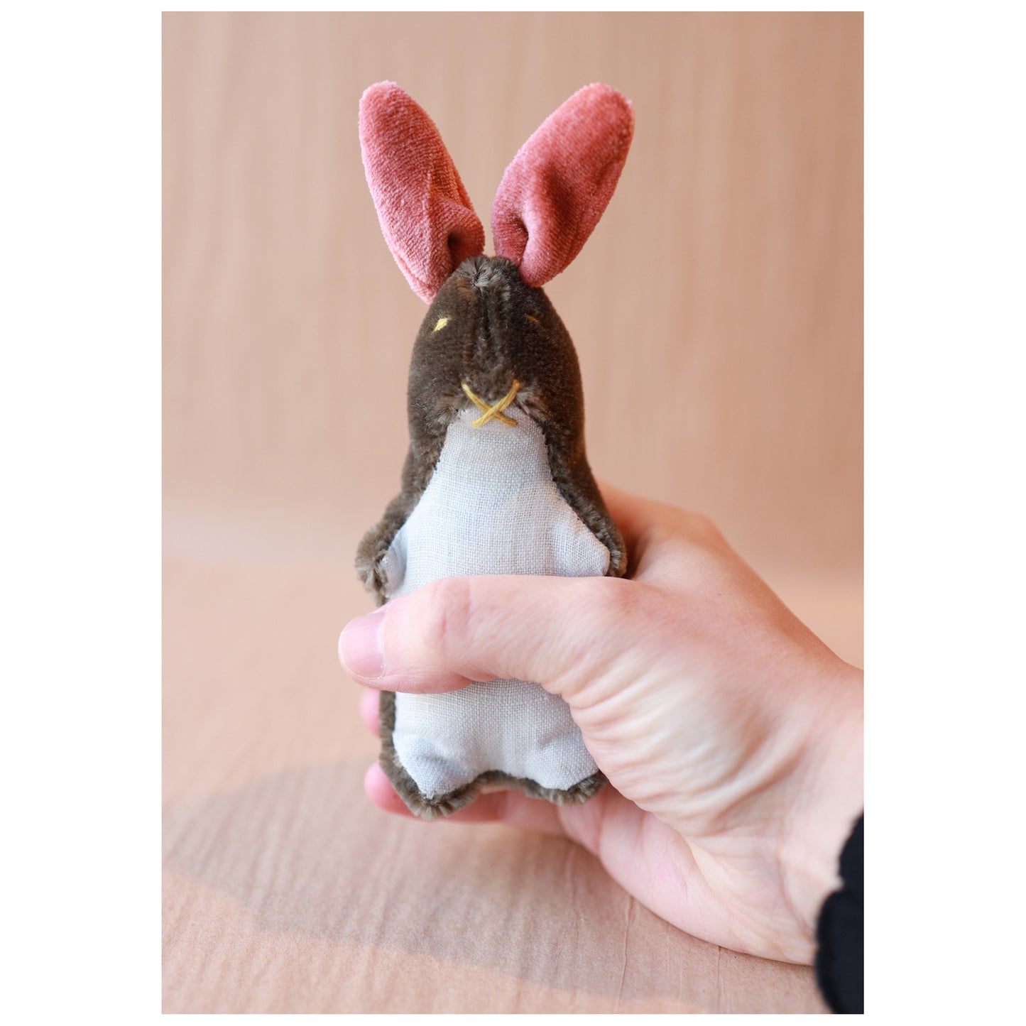 Hand holding a small rabbit-shaped plush toy with pink ears against a beige background