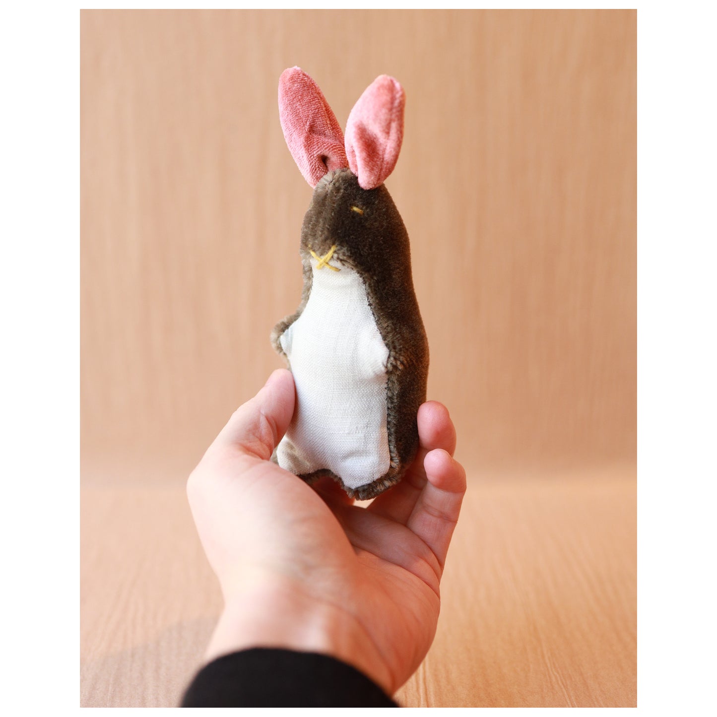 hand holding a small mohair bunny toy with brown mohair, pink ears, and a light blue linen belly