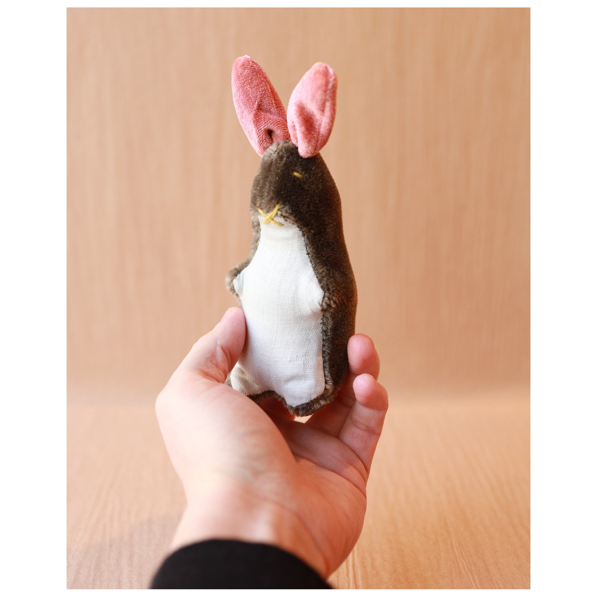 hand holding a small mohair bunny toy with brown mohair, pink ears, and a light blue linen belly