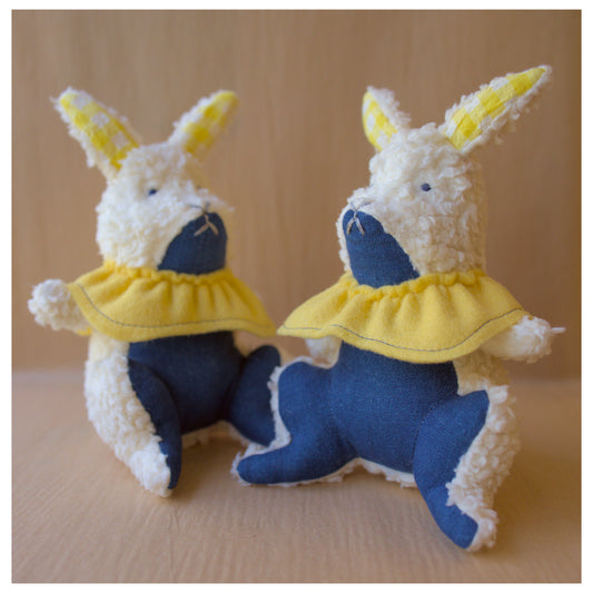 Two plush toys resembling bunnies in blue and yellow outfits on a beige background