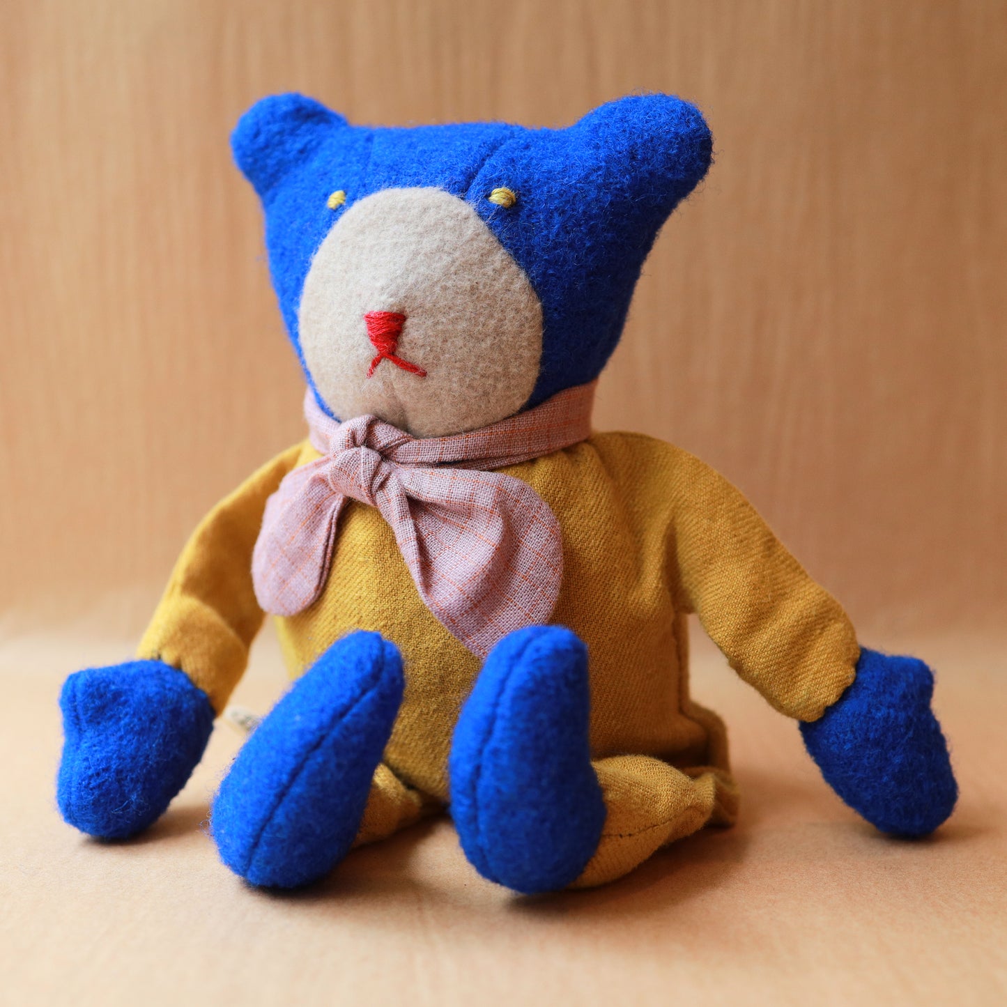 A handmade plush teddy bear made from textured wool-like fabric, featuring a blue head and feet, a mustard yellow body, a light pink bow tie, and a cream-colored snout.