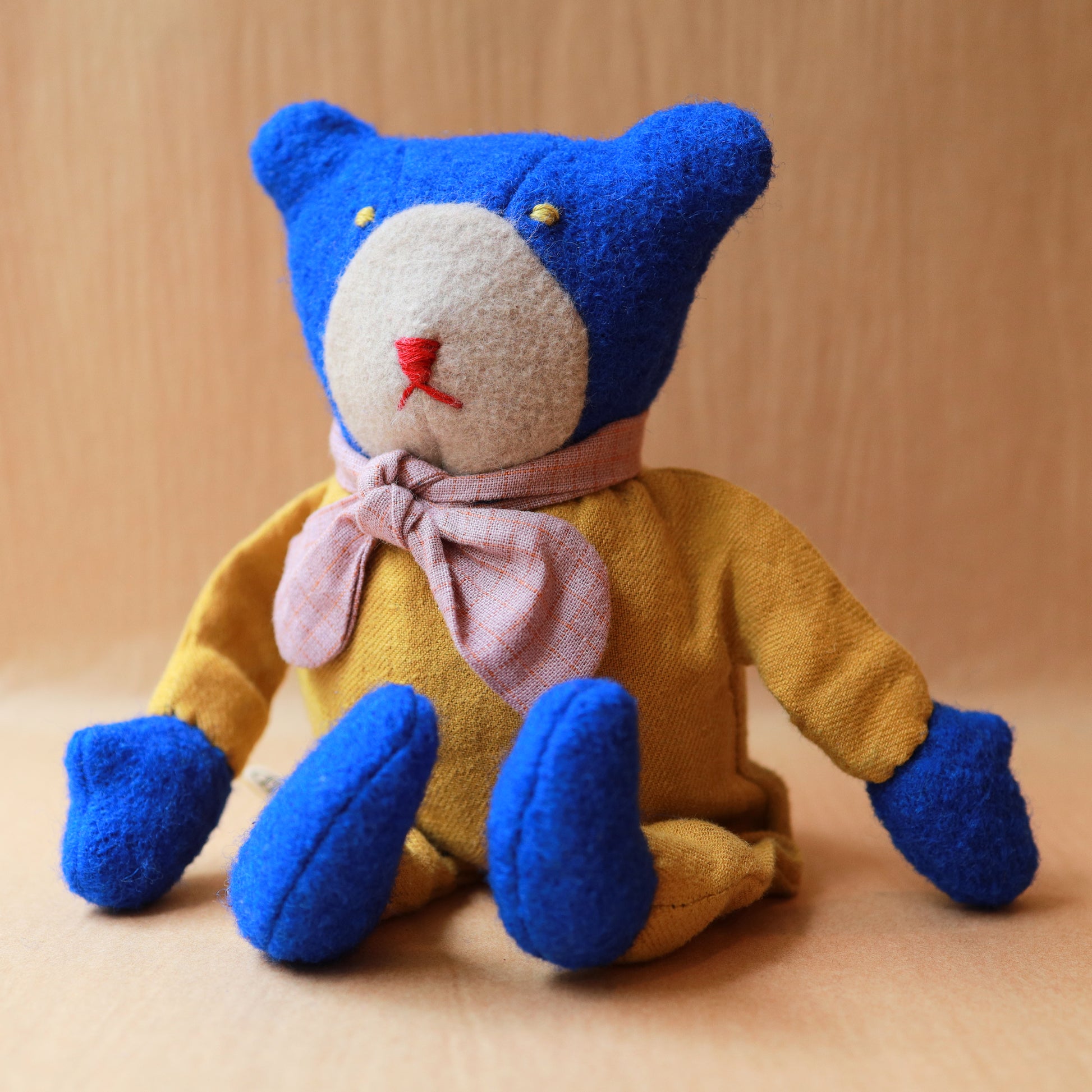 A handmade plush teddy bear made from textured wool-like fabric, featuring a blue head and feet, a mustard yellow body, a light pink bow tie, and a cream-colored snout.