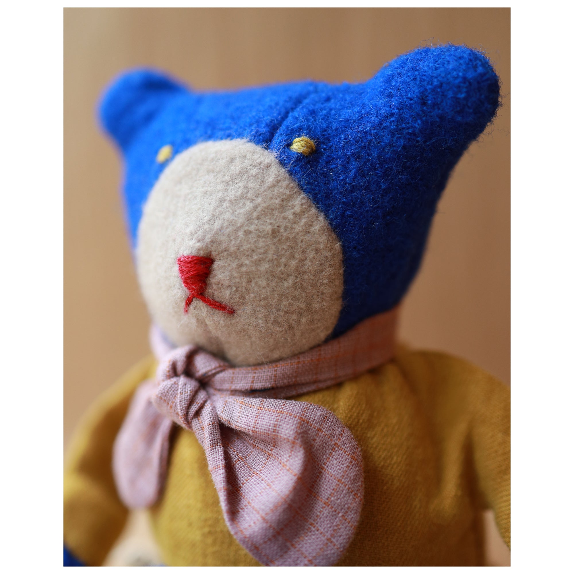 A close-up of a handmade felt teddy bear with a bright blue head, cream-colored snout and ears, a yellow body, and a pink plaid bow tie.