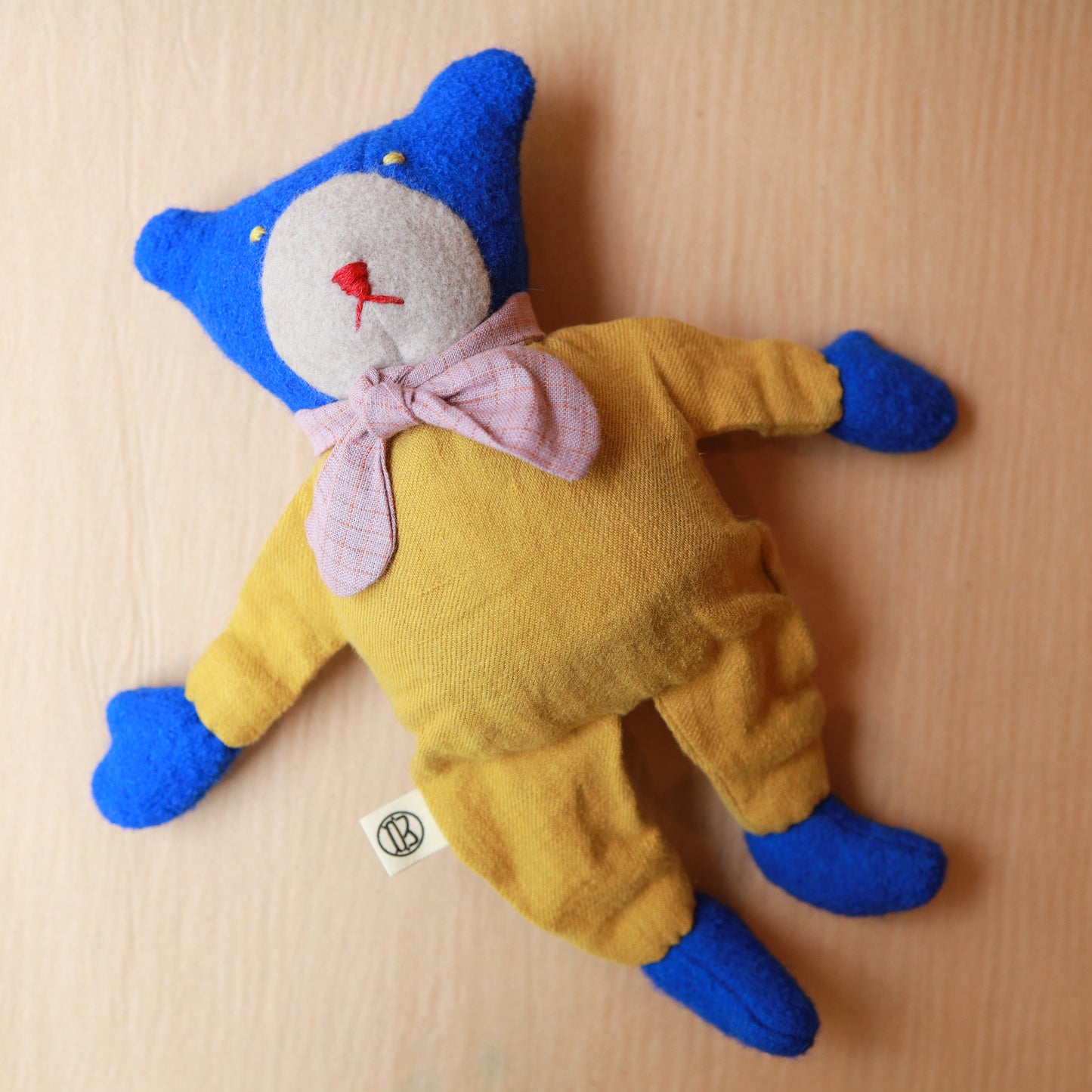 A handmade plush toy bear doll lying flat on a light wood background. The doll has a blue felt head with embroidered eyes and nose, a white face, a pink bow tie, and a yellow fabric body and pants.