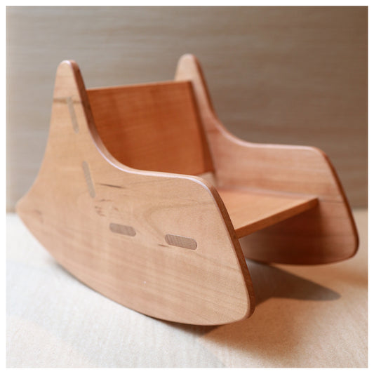 A small wooden rocking chair with a natural light brown finish, photographed on a neutral beige surface against a matching background.