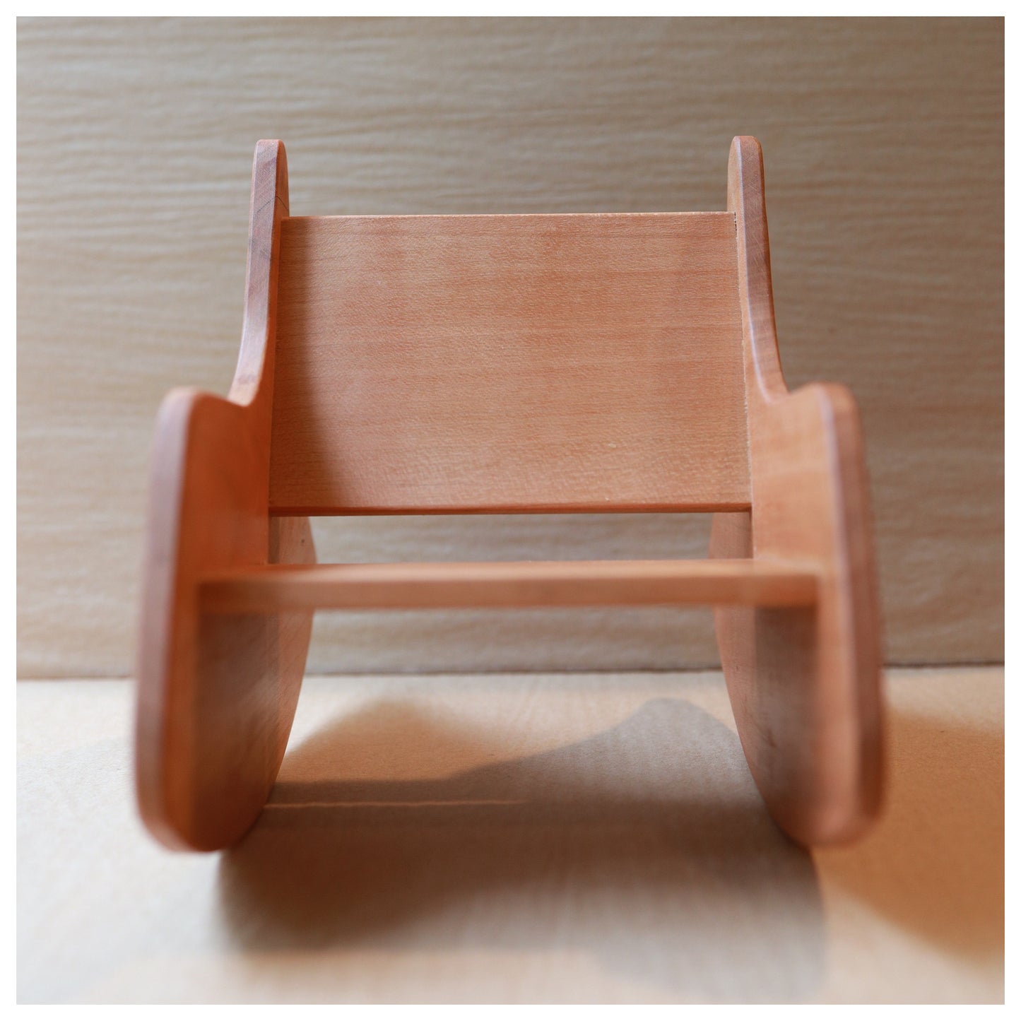 A small wooden rocking toy chair with a natural light brown finish, photographed from a low angle against a light wood background.