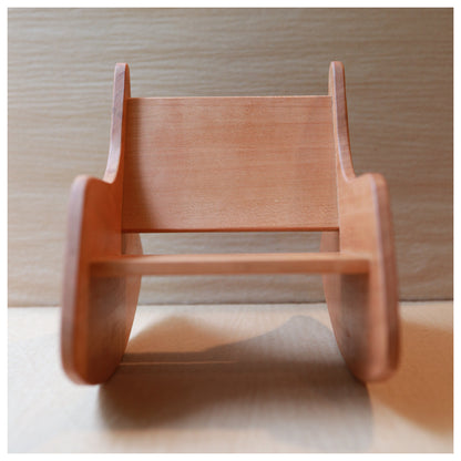 A small wooden rocking toy chair with a natural light brown finish, photographed from a low angle against a light wood background.