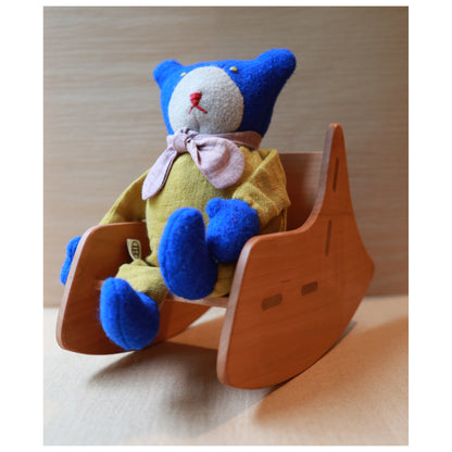 A blue and mustard yellow plush teddy bear wearing a pink bow, sitting in a small wooden rocking chair against a light wood background.