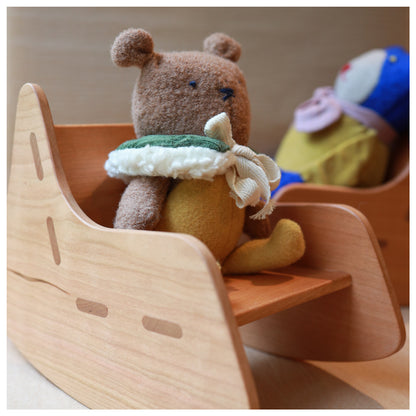 A small plush teddy bear toy wearing a green and cream scarf sits in a wooden rocking chair, with another plush toy blurred in the background.