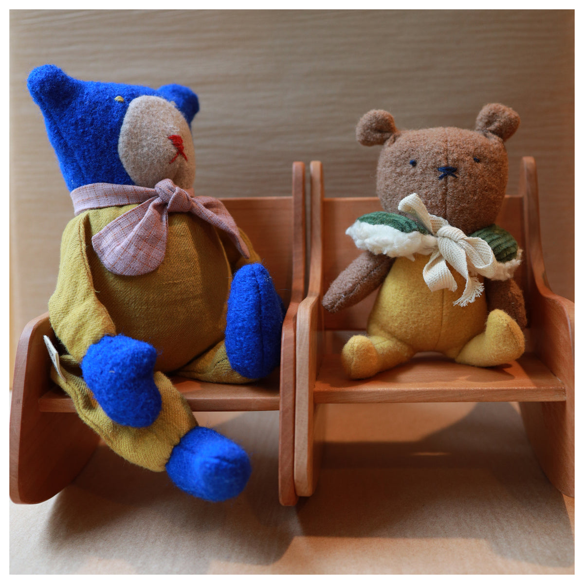 Two small plush teddy bears sitting on miniature wooden chairs against a light wood background.
