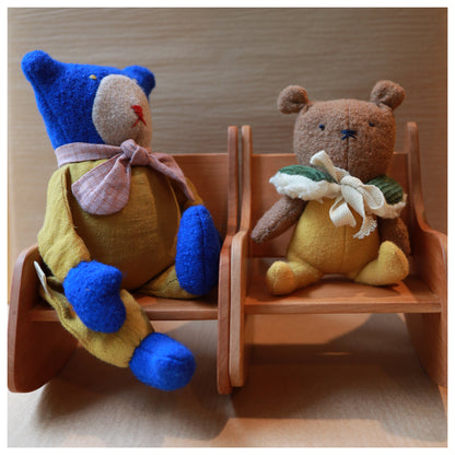 Two small plush teddy bears sitting on miniature wooden chairs against a light wood background.