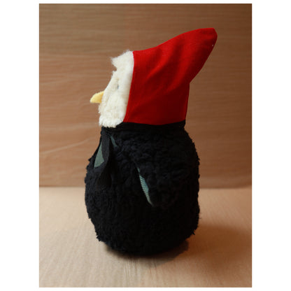 profile of a plush toy owl with a white fluffy face and black body wearing a red hood and a green chest. The beak is yellow felt
