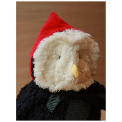 close up face of a plush toy owl with a white fluffy face and black body wearing a red hood and a green chest. The beak is yellow felt