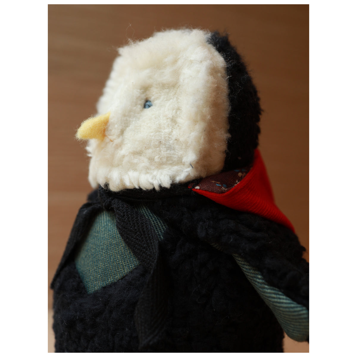 close up with hood down photo of a plush toy owl with a white fluffy face and black body wearing a red hood and a green chest. The beak is yellow felt