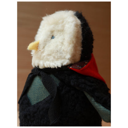 close up with hood down photo of a plush toy owl with a white fluffy face and black body wearing a red hood and a green chest. The beak is yellow felt