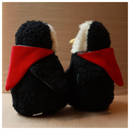 two a plush toy owls with a white fluffy face and black body wearing a red hood and a green chest. The beak is yellow felt