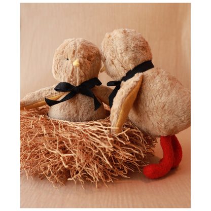 Two plush chicks with black bows sitting on a straw nest against a beige background